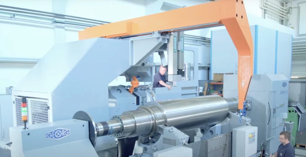 A large industrial lathe machine with an orange overhead arm is operated by a worker in a factory setting.